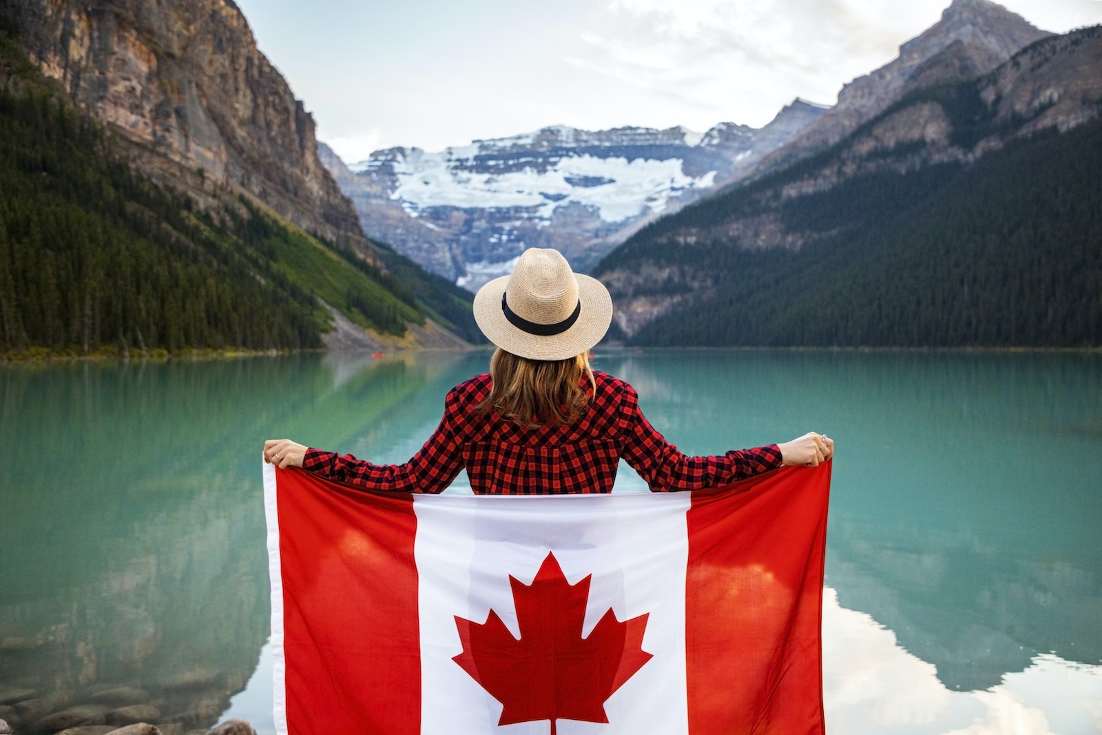 Woman Wearing Red and Black Checkered Dress Shirt and Beige Fedora Hat Holding Canada Flag Looking at Lake